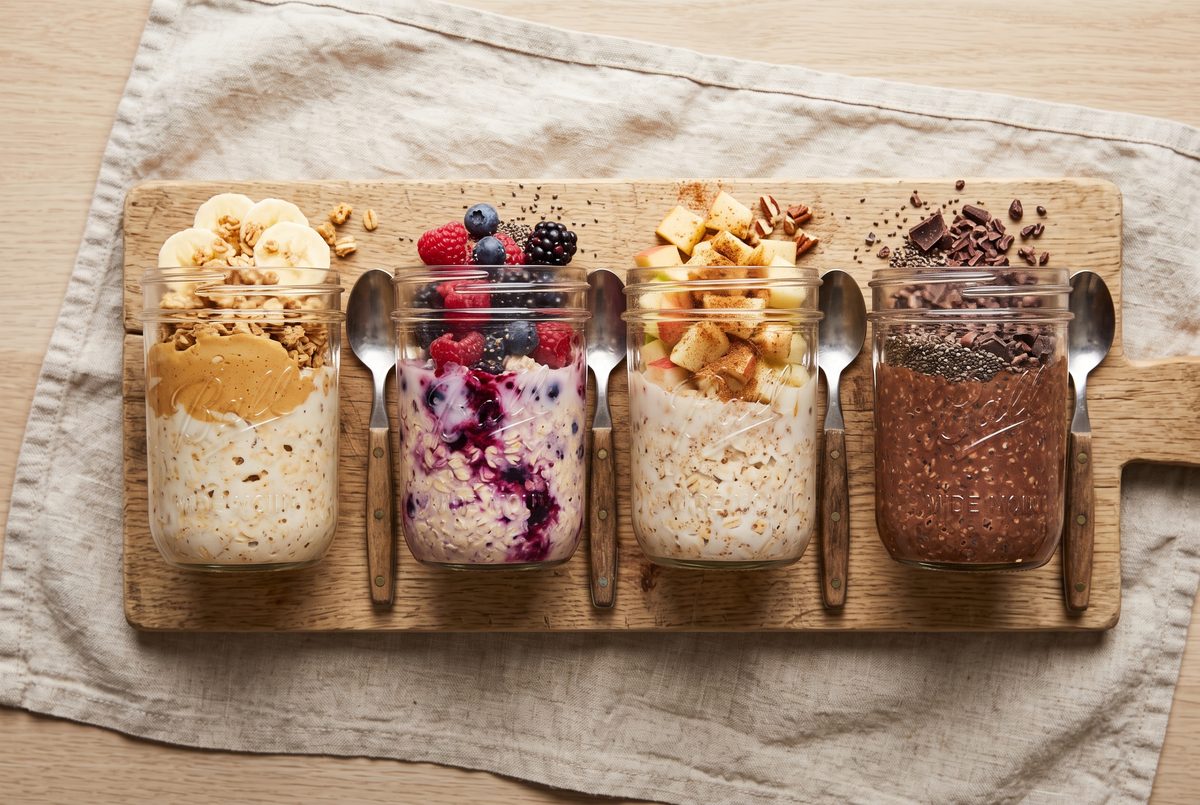 Four glass jars of overnight oats with different toppings — banana and peanut butter, mixed berries, apple cinnamon, and chocolate chia — on a wooden board