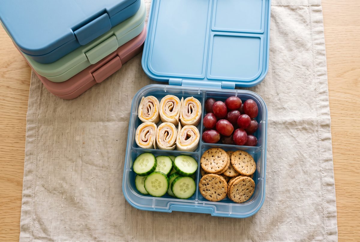 A bento-style lunch box with turkey and cheese tortilla pinwheels, red grapes, cucumber slices and crackers, with three more boxes stacked behind